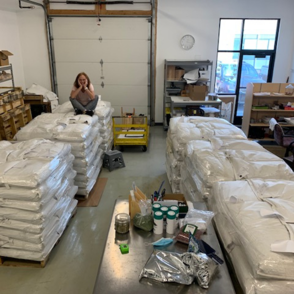 StableFeed owner and founder, Mary Hartman, sitting on a pallet of chia and surrounded by 5 more in her packed store space.